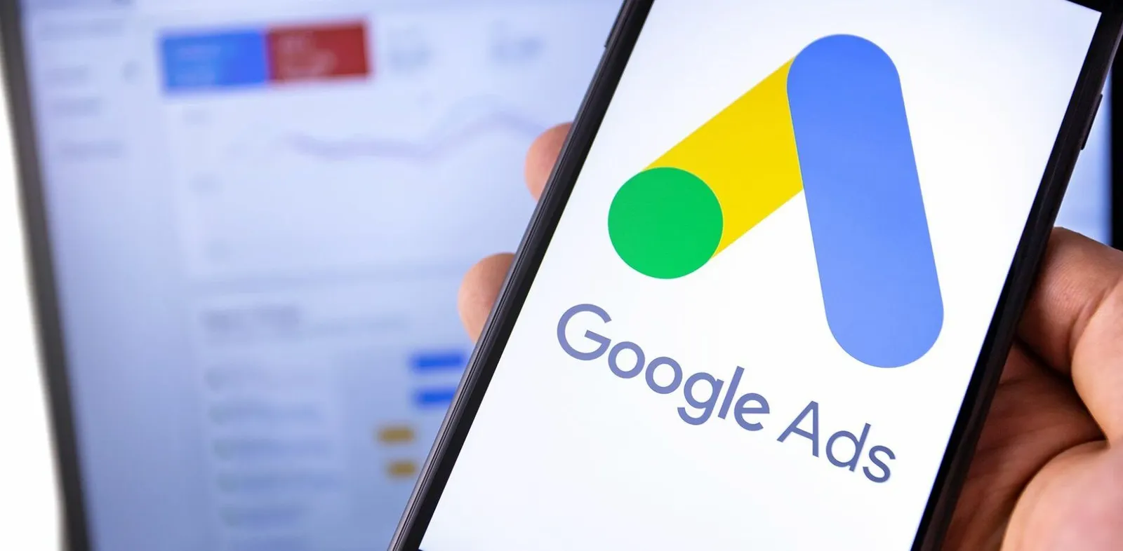 Person holding a phone with the Google Ads logo, with a blurred screen displaying data in the background.