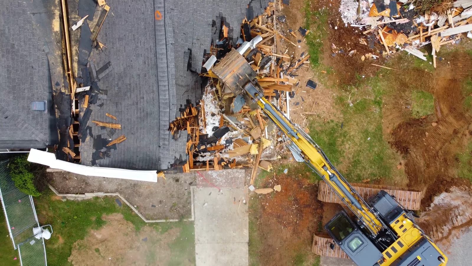 Aerial view of an excavator tearing down a house, roof half gone and debris piled beside it