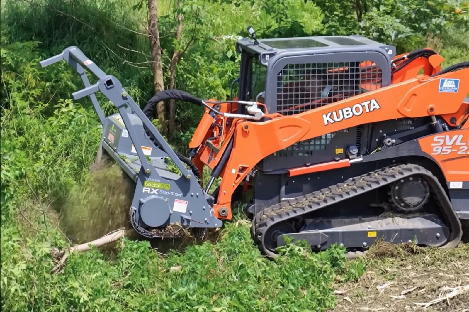 Orange Kubota compact track loader with a forestry mulching head grinding brush on an overgrown lot